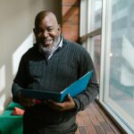 Cheerful man holding a folder in a sunlit room, smiling warmly.